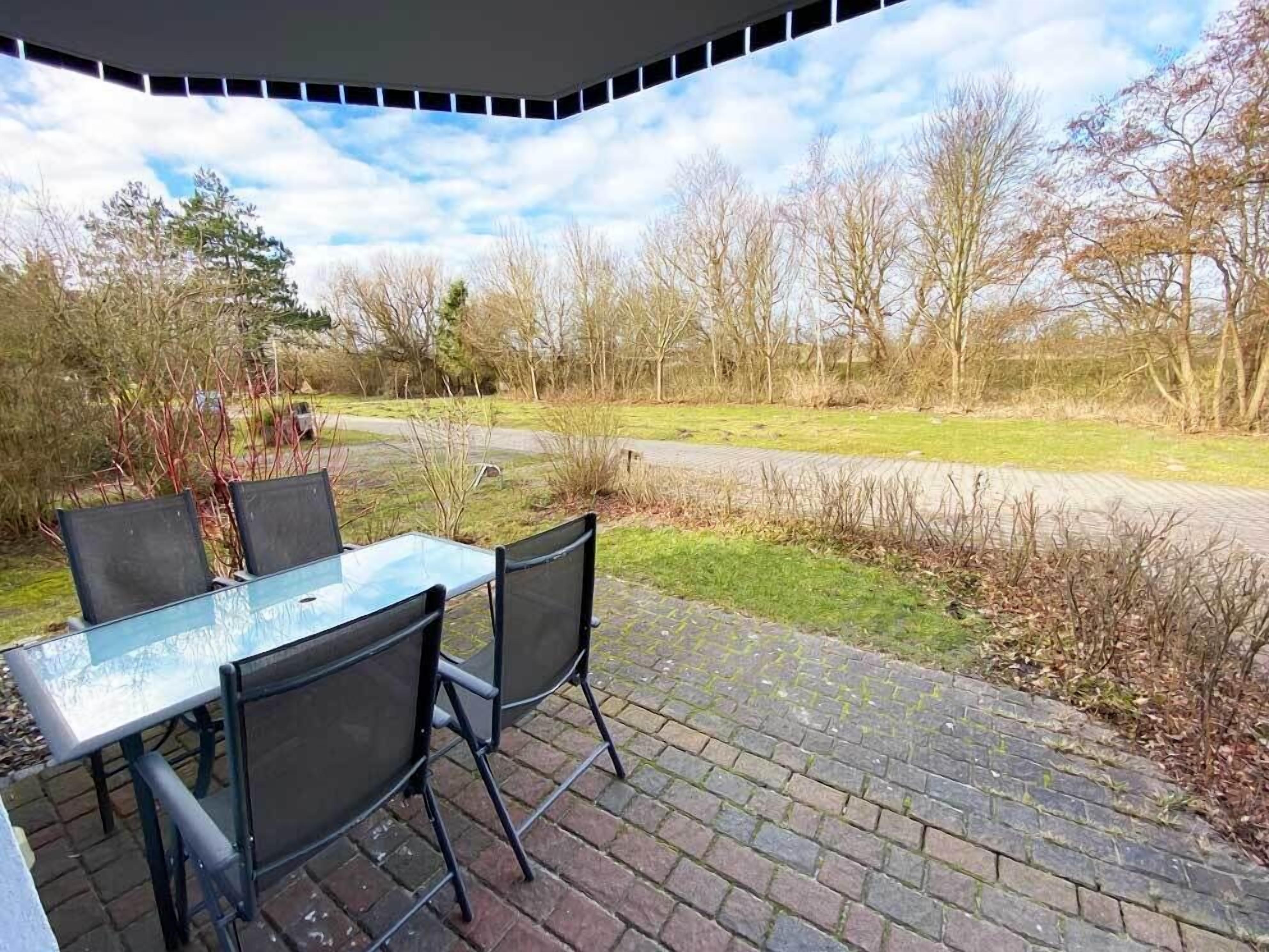An outdoor patio with a glass table and chairs, a brick pathway, and a view of trees and a road.
