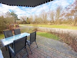 Eine Terrasse im Freien mit einem Glastisch und Stühlen, einem Pflasterweg und Blick auf Bäume und eine Straße.