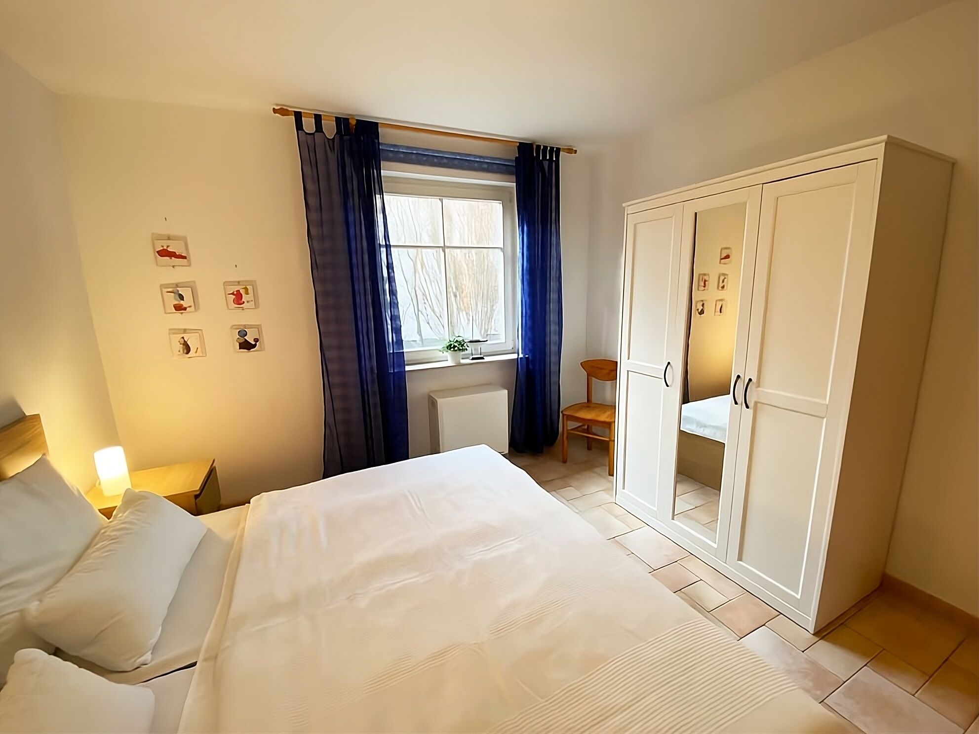 A bedroom with a bed, a wardrobe, a chair, a window with curtains, and wall art.