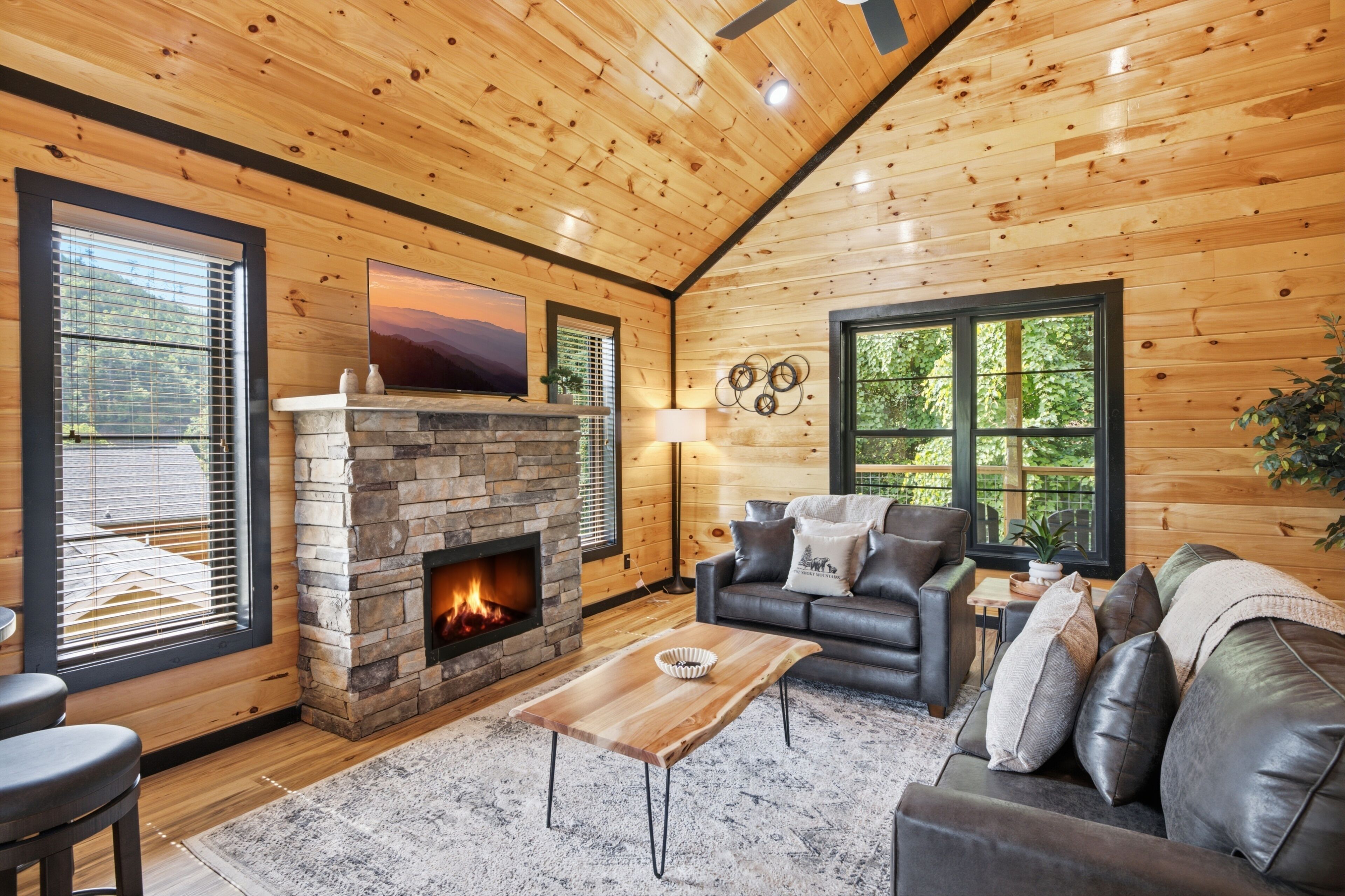A cozy cabin interior with a stone fireplace, leather sofa, and large windows.