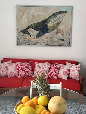 Un canapé rouge avec des coussins à motifs, une table en verre sur laquelle sont disposés des fruits, et un tableau représentant une baleine et la Tour Eiffel.