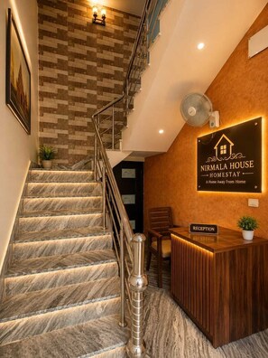 Ein Empfangsschalter mit dem Schriftzug 'Nirmala House Homestay', eine Treppe mit Marmorstufen und eine Wand mit Fischgrätmuster.