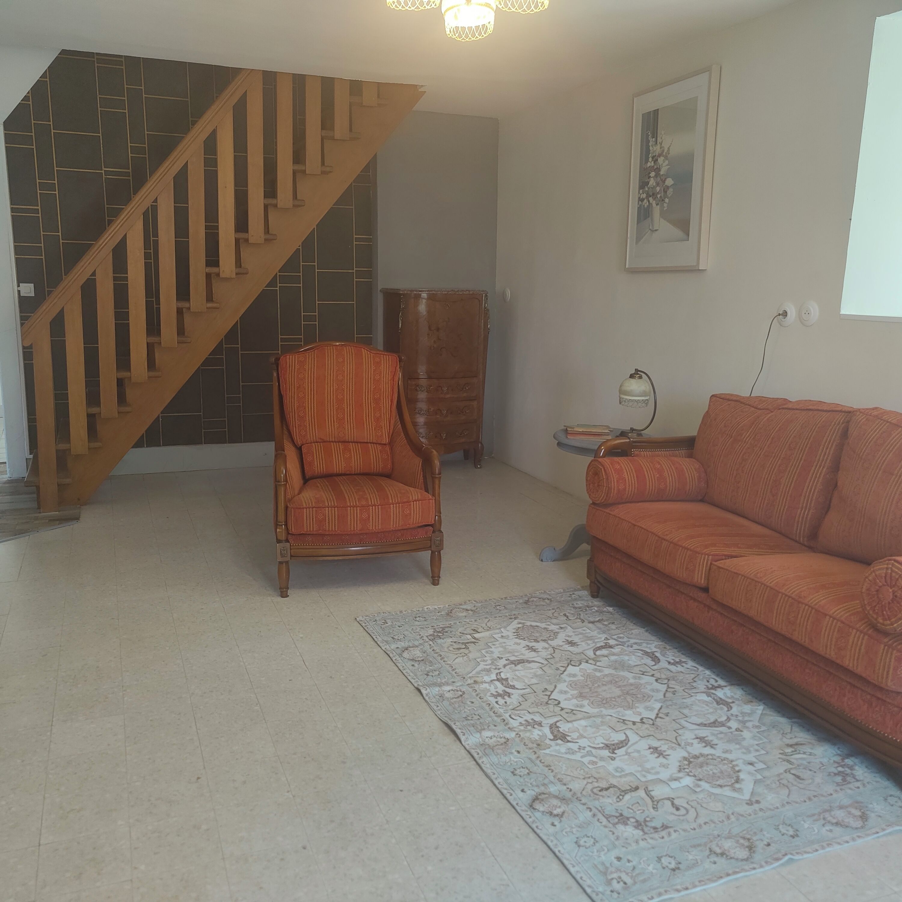 A living room with a wooden staircase, a patterned armchair, a sofa, a cabinet, and a rug.