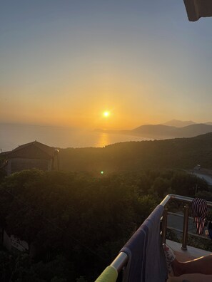 Vista del atardecer desde un balcón con vista al agua, con montañas a lo lejos y una casa con techo de tejas.