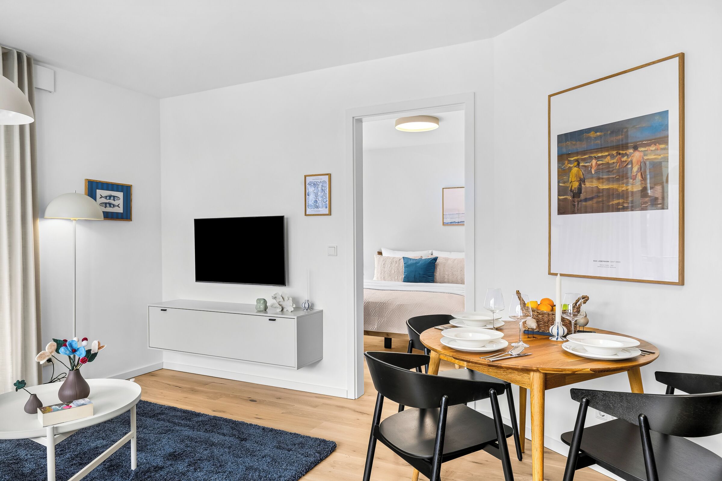 Une chambre d’hôtel moderne équipée d’un téléviseur à écran plat, d’une table à manger dressée pour deux personnes et d’un lit avec un oreiller bleu.