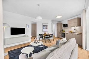 A modern living room with a flat-screen TV, a grey sofa, a dining table with chairs, and a kitchen area with cabinets and a sink. - The View A04 Cloudsurfer  – Balcony, Garden Furniture, Shared Sauna, Parking (Dagebüll)