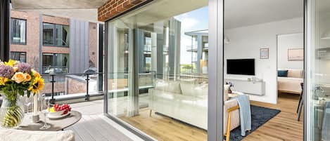 A modern apartment with a balcony, a dining area with a table and chairs, and a living room with a sofa and a TV.