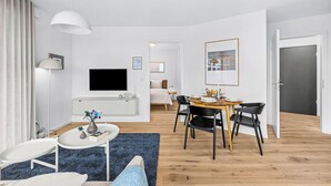 A modern living room with a flat-screen TV, a dining area with a wooden table and black chairs, and a hallway leading to a bedroom. - The View A04 Cloudsurfer  – Balcony, Garden Furniture, Shared Sauna, Parking (Dagebüll)