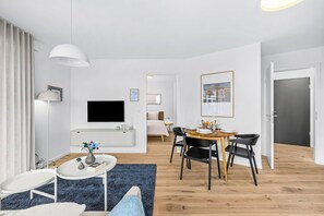 Ein modernes Wohnzimmer mit Flachbildfernseher, eine Essecke mit Holztisch und schwarzen Stühlen sowie ein Flur, der zum Schlafzimmer führt.