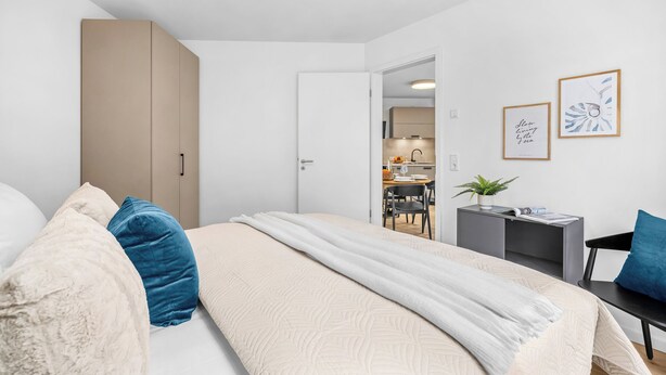 A modern bedroom with a bed, a desk, and a kitchenette in the background. - The View A04 Cloudsurfer  – Balcony, Garden Furniture, Shared Sauna, Parking (Dagebüll)