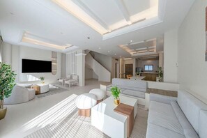 A modern living room with a flat-screen TV, white sofas, a coffee table, and a staircase leading to another level. - Mabat Jasmine Greek Villa 4BR 583 Villa6 (Jeddah)