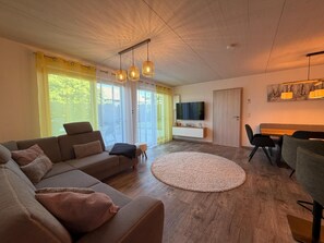 A modern living room with a sofa, a round rug, a dining area with chairs, a TV mounted on the wall, and large windows with curtains. - Time out by the lake: Luxurious vacation apartment with garden (Gaienhofen)