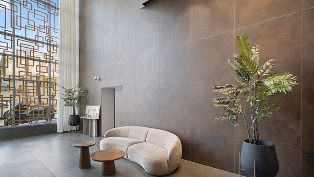 Un vestíbulo moderno con un sofá curvo, dos mesas redondas de madera, una planta grande en maceta y vistas al exterior a través de una ventana con diseño.