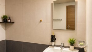 A bathroom with a white sink, a mirror, a wall-mounted light, and a shelf with toiletries.