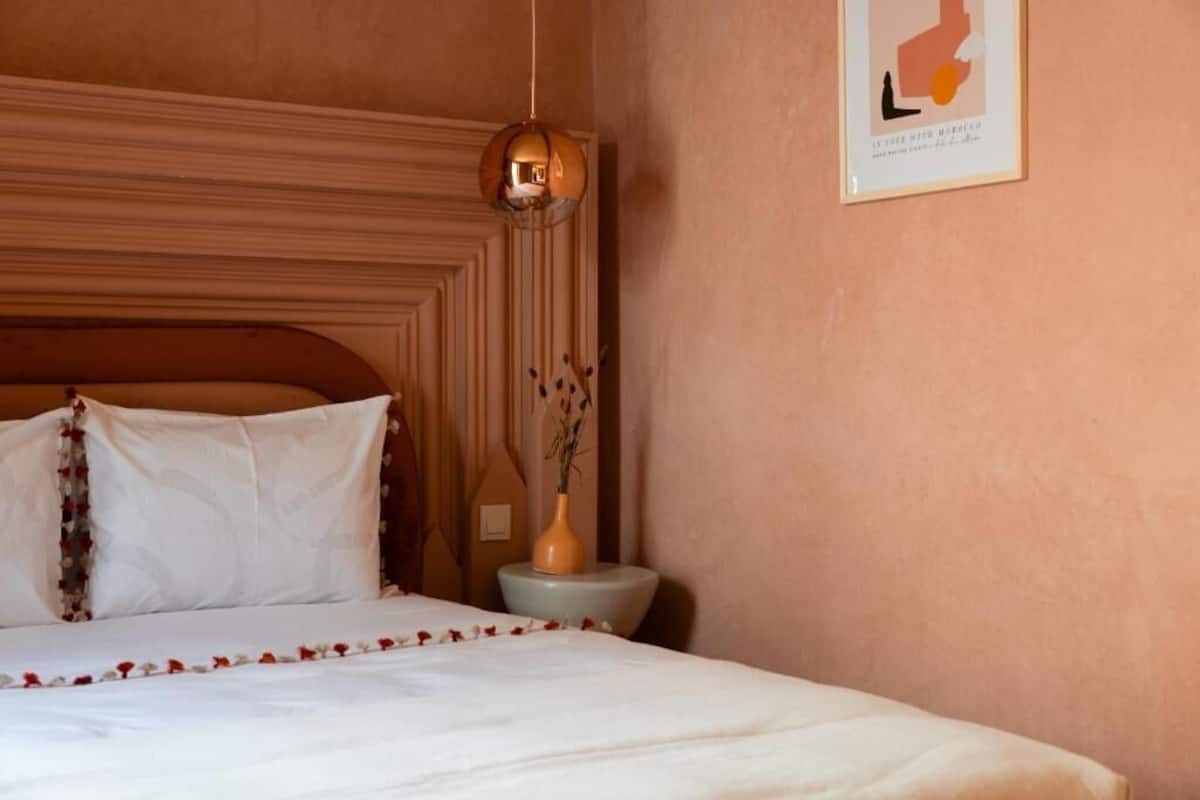 Un lit bien fait, avec du linge de lit blanc et un motif décoratif sur les bords, un vase contenant des fleurs séchées, un luminaire fixé au mur et une carte encadrée accrochée au mur.