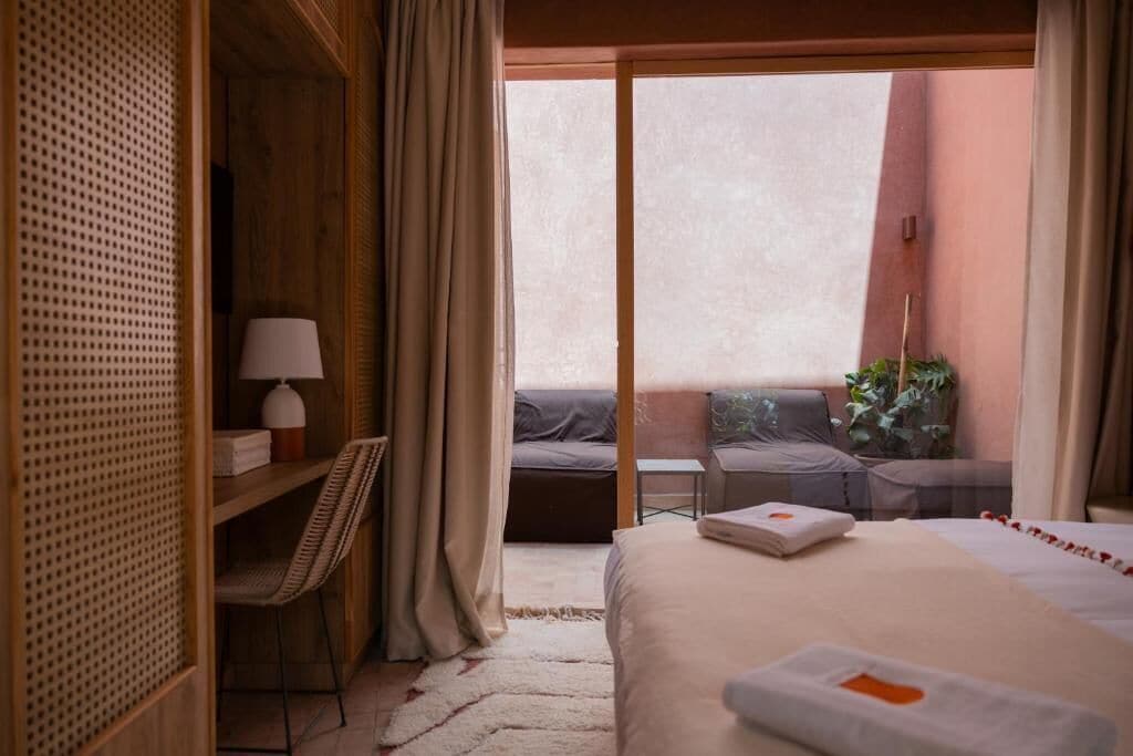A hotel room with a bed, a desk with a lamp, a chair, a small table, and a view of an outdoor seating area.