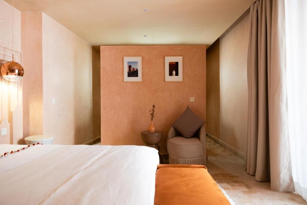 A hotel room with a bed, a chair, a small table, and two framed pictures on the wall.