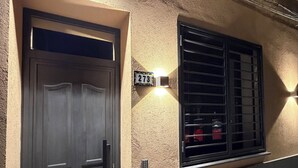 Fachada de edificio con una puerta de madera oscura, una ventana con barrotes y una lámpara fijada a la pared.