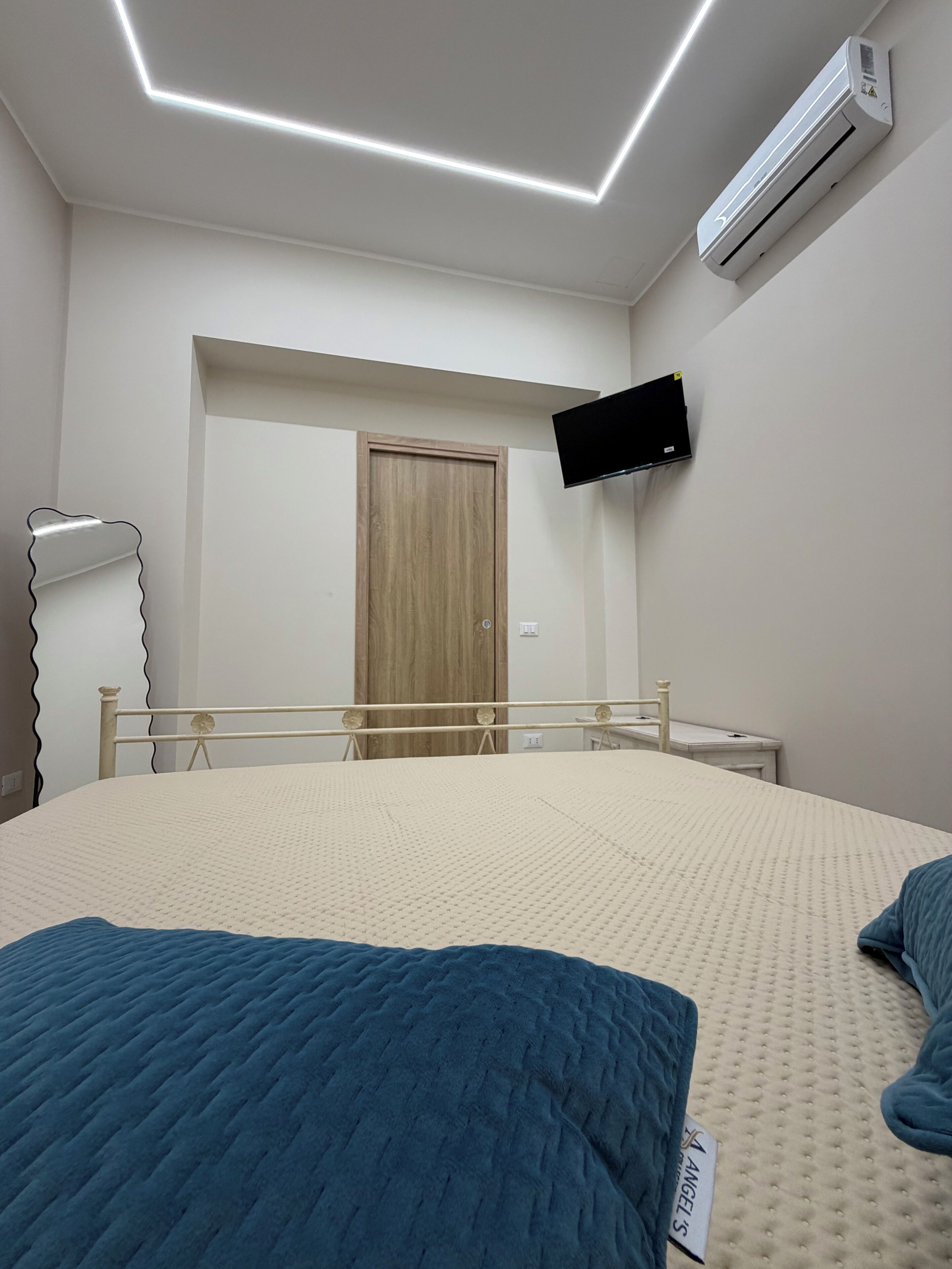 A bedroom with a bed, a television mounted on the wall, an air conditioner, a wooden door, and a bedside table.