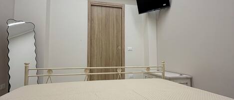 A bedroom with a bed, a television mounted on the wall, an air conditioner, a wooden door, and a bedside table.