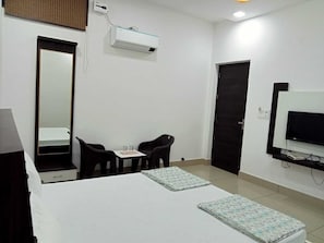 A hotel room with a bed, a desk with two chairs, a TV, and an air conditioning unit.