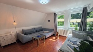 Ein modernes Wohnzimmer mit einem grauen Sofa, einem Glastisch, einer weißen Kommode und einem großen Fenster mit Vorhängen.