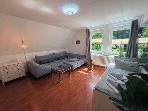 Ein modernes Wohnzimmer mit einem grauen Sofa, einem Glastisch, einer weißen Kommode und einem großen Fenster mit Vorhängen.