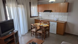 Um espaço de estar compacto com televisão, mesa de jantar, cadeiras, sofá, cozinha com armários e máquina de lavar loiça, e uma janela com cortinas.