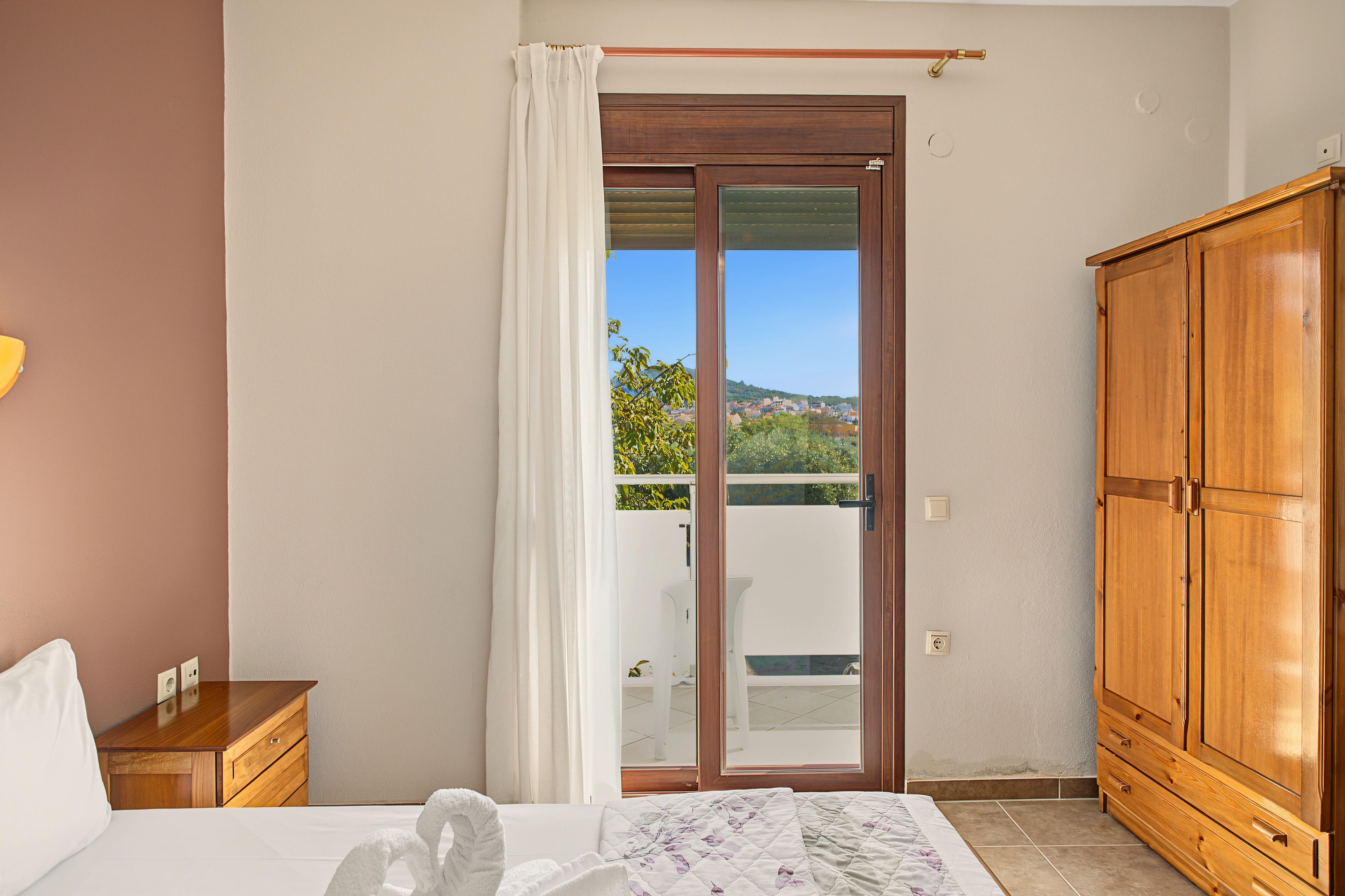 Ein Schlafzimmer mit einem Bett, einem Holzschrank, einem Nachttisch und einer Schiebetür zu einem Balkon mit Blick auf Gebäude und Grünflächen.