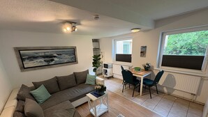 Ein modernes Wohnzimmer mit einer Eckcouch, einem Esstisch mit Stühlen, einer Küchenzeile und einem großen Fenster mit Blick auf Grünflächen.