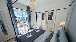 Ein Bett mit Baldachin, ausgestattet mit einem dunklen Bettbezug und Kissen, eine Kristallleuchter, ein weißer Kleiderschrank und ein Blick ins Freie.