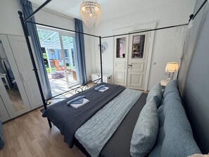 Ein Bett mit Baldachin, ausgestattet mit einem dunklen Bettbezug und Kissen, eine Kristallleuchter, ein weißer Kleiderschrank und ein Blick ins Freie.