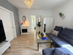 Ein Wohnzimmer mit Sofa, Glastisch, Kamin, Fernseher und einer Chandelüster.