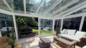 Ein sonnendurchfluteter Raum mit Glasdach, einem weißen Sofa, einem runden Hocker, einem Korbstuhl und Blick auf einen Garten.