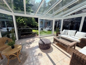 Ein sonnendurchfluteter Raum mit Glasdach, einem weißen Sofa, einem runden Hocker, einem Korbstuhl und Blick auf einen Garten.