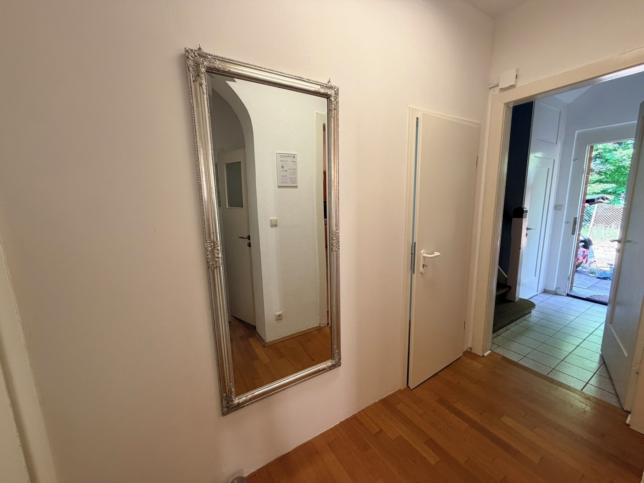 Een gang met een grote spiegel, een deur en een zicht op een kamer met betegelde vloer.