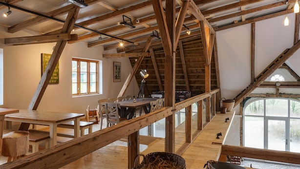 Ein rustikaler Innenraum mit freiliegenden Holzbalken, einem Essbereich mit Tischen und Stühlen sowie einem großen Fenster, das natürliches Licht hereinlässt.