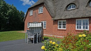 Ein rotes Backsteinhaus mit einem Reetdach, einer Terrasse mit Tisch und Stühlen und einem Garten mit gelben Blumen.