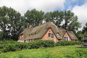 Ein traditionelles Haus mit Strohdach und Ziegelsohle, umgeben von üppigem Grün und Bäumen.