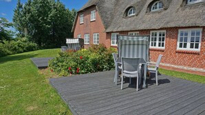 Ein Haus mit Strohdach, Ziegelaußenwand, einer Holzterrasse und einem Garten mit roten Blumen.