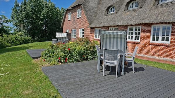 Ein Haus mit Strohdach, Ziegelaußenwand, einer Holzterrasse und einem Garten mit roten Blumen.