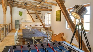 Ein Zimmer mit Holzboden, einem Kicker, einem Esstisch mit Stühlen, einer Küchenzeile und einem Fenster mit Tageslicht.