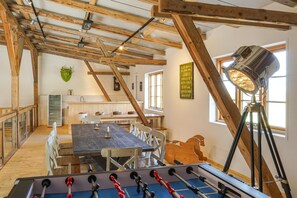 Una habitación con techo de madera, mesa de futbolín, mesa de comedor con sillas, zona de cocina y ventana con luz natural.