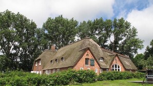 Ein traditionelles Haus mit Strohdach und Ziegelsohle, umgeben von üppigem Grün und Bäumen.