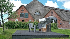 Ein Backsteinhaus mit einem Reetdach, ein kleiner Garten mit Stühlen und einem Tisch, und ein klarer Himmel.