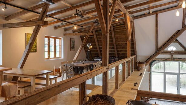 Ein rustikaler Innenraum mit freiliegenden Holzbalken, einem Essbereich mit Tischen und Stühlen sowie einem großen Fenster, das natürliches Licht hereinlässt.