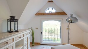 Ein Schlafzimmer mit schrägen Wänden, einem Bett, einem Schreibtisch mit Schubladen, einer Lampe und einem Fenster mit Blick ins Freie.