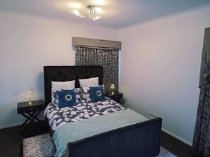 A bedroom with a bed, two bedside tables, a chandelier, a window with curtains, and a patterned rug. - Panoramic View in Narre Warren North (Melbourne)