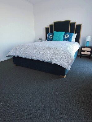 A bed with a patterned bedspread, a headboard with a decorative panel, a bedside table with a lamp, and a carpeted floor.