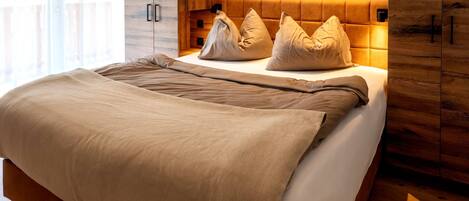 A modern bedroom with a wooden headboard, a large bed with beige bedding, and a view of a forest through the window.